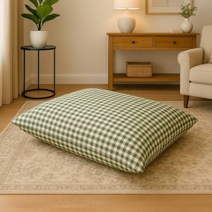 May include: A large, square floor cushion with a green and white gingham pattern. The cushion is placed on a patterned rug in a living room setting. The cushion is a soft, comfortable addition to any room.