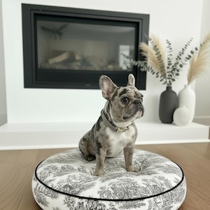 May include: A French Bulldog with a gray and white coat sits on a round dog bed. The bed has a black and white toile pattern and a black trim. The dog wears a gray collar with a gold buckle. The background includes a fireplace and vases.