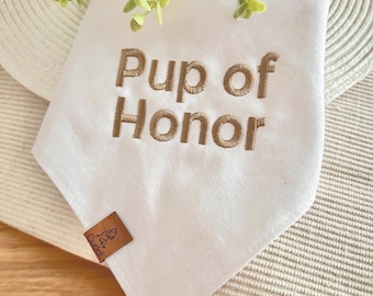 Peut inclure: Bandana blanche pour chien avec l'inscription brodée "Pup of Honor" en fil marron. Une petite étiquette en cuir marron avec un logo est attachée. Le bandana est plié sur une surface tissée avec de la verdure en arrière-plan.