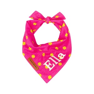 May include: Pink dog bandana with a yellow softball pattern and the name "Ella" embroidered in white. The bandana is tied in a knot at the top and has a small brown tag that says "Sammy's Sew Shop".