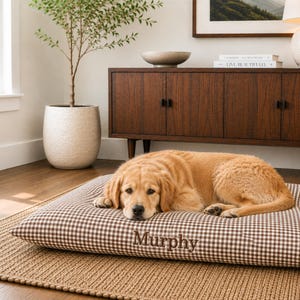 Puede incluir: Una cama rectangular para mascotas con un patrón de cuadros vichy en marrón y blanco. La tela está bordada con el nombre Murphy en una fuente cursiva de color marrón oscuro. Un perro de color dorado está acostado en el centro de la cama.
