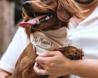 Peut inclure: Un chien roux heureux portant un bandana beige avec l'inscription dorée "The Mason's Est. 2026". Le chien est tenu par une personne, et une bague en diamant est visible. La langue du chien est sortie. L'image suggère un mariage ou des fiançailles.