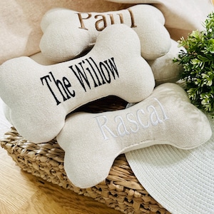 May include: Several beige, bone-shaped dog toys with embroidered names: "Paul", "The Willow", and "Rascal". The toys are made of a textured fabric and are arranged in a woven basket, with greenery in the background.