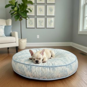 May include: A sleeping fawn-colored French Bulldog rests on a round dog bed. The bed features a light blue and white toile pattern with a matching blue trim. The bed is on a hardwood floor.