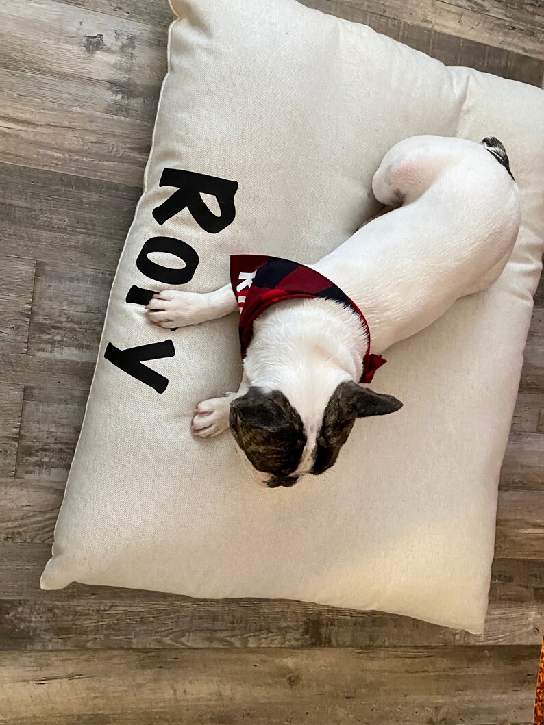May include: A white dog bed with the name "ROLY" printed in black on the side. A white and brown dog with a red and black bandana is lying on the bed.