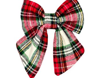 May include: A large, fabric bow with a classic plaid pattern in red, green, white, and yellow. The bow is symmetrical, with two large loops and two long, pointed tails. The fabric has a textured appearance.