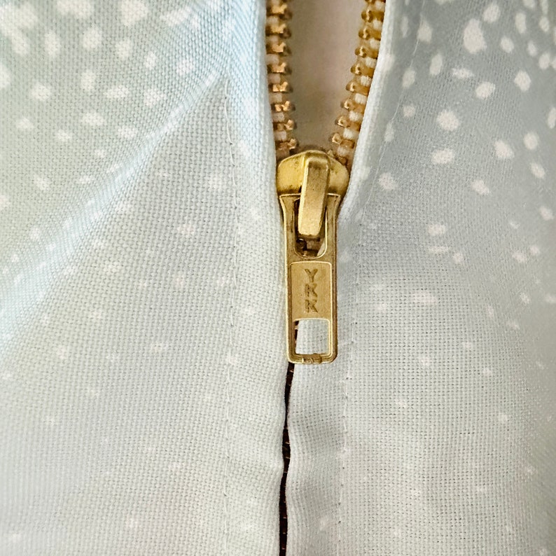 May include: Close-up of a gold zipper with the text "YKK" on the pull tab. The zipper is sewn into a light blue fabric with a white polka dot pattern.