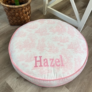 Puede incluir: Un cojín de suelo redondo de color rosa y blanco con estampado y un borde rosa con el nombre "Hazel" bordado en rosa.