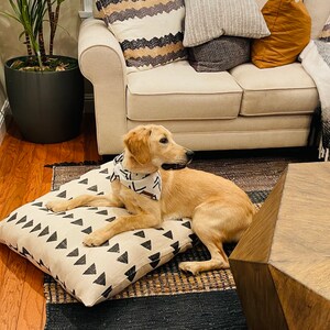 May include: A light brown dog wearing a white and black bandana is lying on a white and black patterned dog bed. The dog is looking to the right of the image. The dog bed is on a brown and black rug. There is a wooden coffee table in the background.