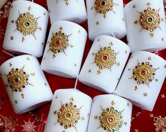 May include: A collection of 10 white pillar candles decorated with gold and colorful rhinestones. The candles are arranged on a red plate with a decorative pattern.