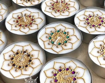 May include: Assortment of white tea light candles decorated with gold henna designs and colorful rhinestones. The candles are arranged in a circular pattern on a wooden surface. The text "CREATIVE HENNA CRAFTS" is visible at the bottom of the image.