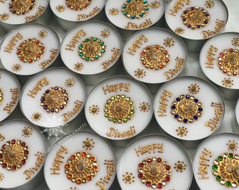 May include: A collection of white tea light candles, each featuring the gold-colored text Happy Diwali and a central circular gold filigree ornament decorated with small, colorful rhinestones in various hues surrounding the candle wick.