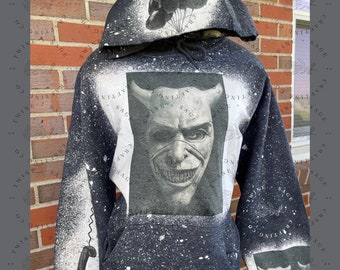 May include: A black hooded sweatshirt with a bleached splatter effect. The front features a large rectangular graphic of a horned, smiling face. The hood displays a cluster of balloons, and the sleeve shows a telephone receiver graphic.