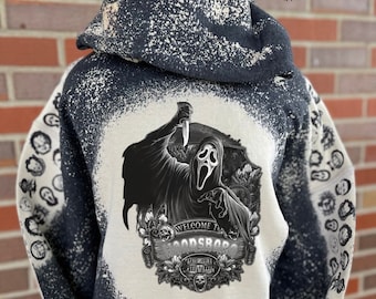 May include: A black hoodie with a bleached, mottled pattern featuring a graphic of a masked figure holding a knife above the text Welcome to Woodsboro. The sleeves display a repeating pattern of small masked faces, and the back features a large central print.