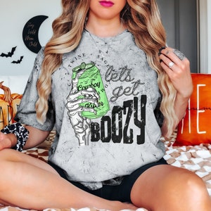 May include: Gray tie-dye t-shirt with a graphic of a skeleton hand holding a green can of "Cowboy Water". The text "lets get BOOZY" is printed in black. The shirt has a crew neck and short sleeves.