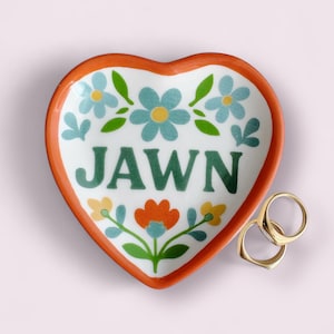 May include: Heart-shaped ceramic trinket dish with an orange rim and floral design. The word "JAWN" is written in green. Two gold rings are next to the dish. The background is a light pink.