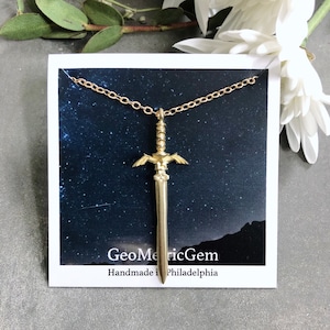 Può includere: Una collana d'oro con un ciondolo a forma di spada. La spada ha le ali ed è dettagliata. La collana è su una carta con uno sfondo stellato e il testo "GeometricGem Handmade in Philadelphia".