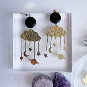 May include: A pair of celestial-themed earrings. Each earring features a black circular top, a textured gold-colored cloud, and dangling charms of stars, moons, and a planet. The earrings are displayed on a clear acrylic block.