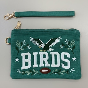 May include: A teal canvas zipped pouch with a detachable wrist strap. The front features an embroidered bald eagle, the word BIRDS in white block letters, a brown football, white stars, and green leafy branches with gold-toned hardware.