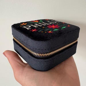 May include: A black, square jewellery box with a gold zip, held in a hand. The top is embroidered with the name "PHILL" in white, surrounded by red and orange flowers with green leaves. The box is made of a soft, velvety material.