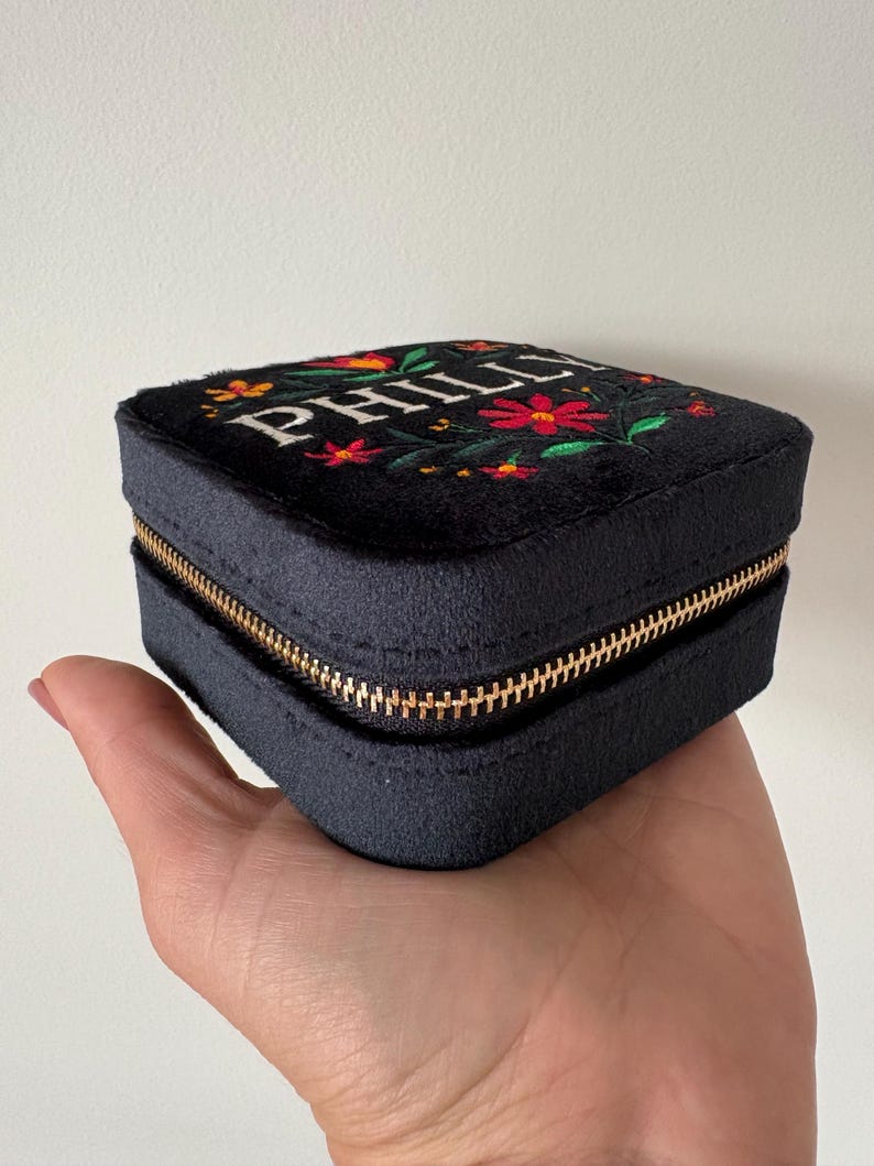 May include: A black, square jewellery box with a gold zip, held in a hand. The top is embroidered with the name "PHILL" in white, surrounded by red and orange flowers with green leaves. The box is made of a soft, velvety material.