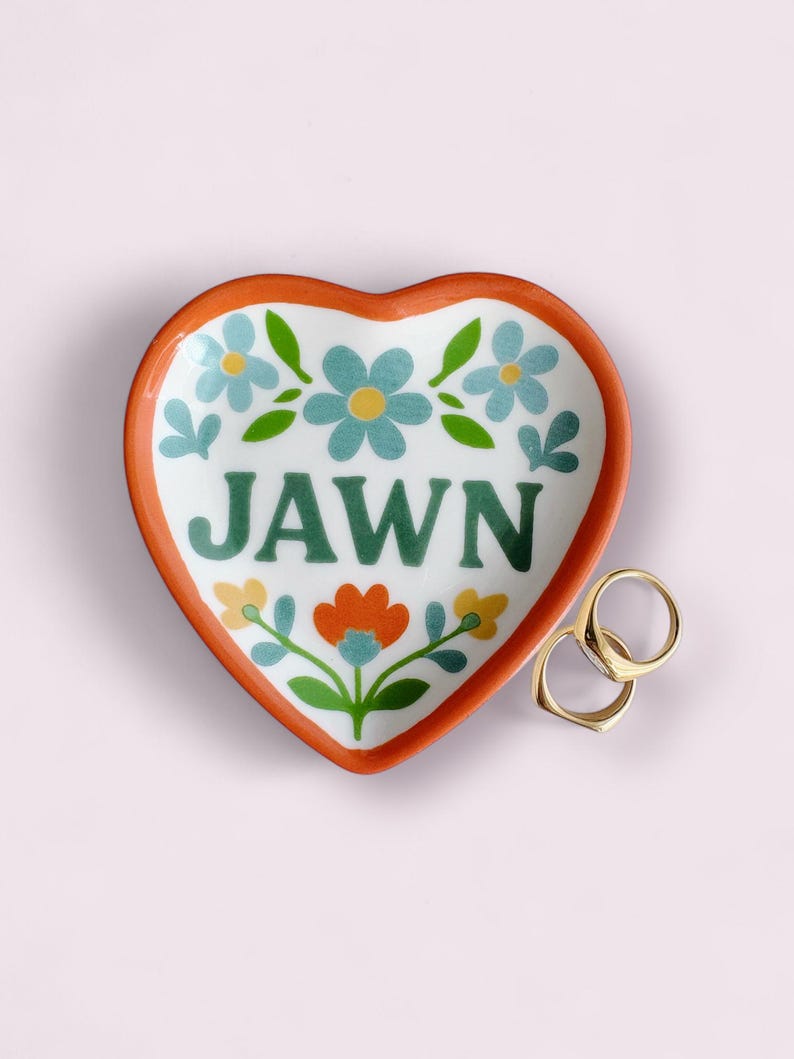 Op de afbeelding: Hartvormig keramisch sieradenschaaltje met een oranje rand en bloemenmotief. Het woord "JAWN" is in het groen geschreven. Twee gouden ringen liggen naast het schaaltje. De achtergrond is lichtroze.