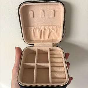 May include: Open black jewellery travel case with a gold zip and a beige interior. The interior has compartments for rings, earrings, and other jewellery. The case is held open by a hand.