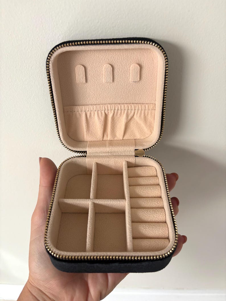 May include: Open black jewellery travel case with a gold zip and a beige interior. The interior has compartments for rings, earrings, and other jewellery. The case is held open by a hand.