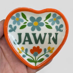 Op de afbeelding: Hartvormige keramische schaal met een oranje rand en bloemmotief. De schaal heeft blauwe en gele bloemen, groene bladeren en het woord "JAWN" in groene blokletters. Een decoratief stuk voor in huis.