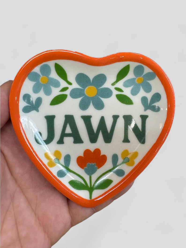 Op de afbeelding: Hartvormige keramische schaal met een oranje rand en bloemmotief. De schaal heeft blauwe en gele bloemen, groene bladeren en het woord "JAWN" in groene blokletters. Een decoratief stuk voor in huis.
