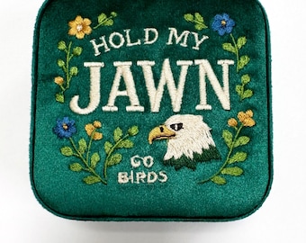 May include: A square, emerald green velvet jewelry box with embroidered text and floral accents. The text reads "HOLD MY JAWN" above an eagle head and the words "GO BIRDS". The embroidery includes yellow and blue flowers.