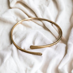 May include: A simple gold choker necklace with a rounded shape and a slightly angled end. The necklace is lying on a white fabric background.