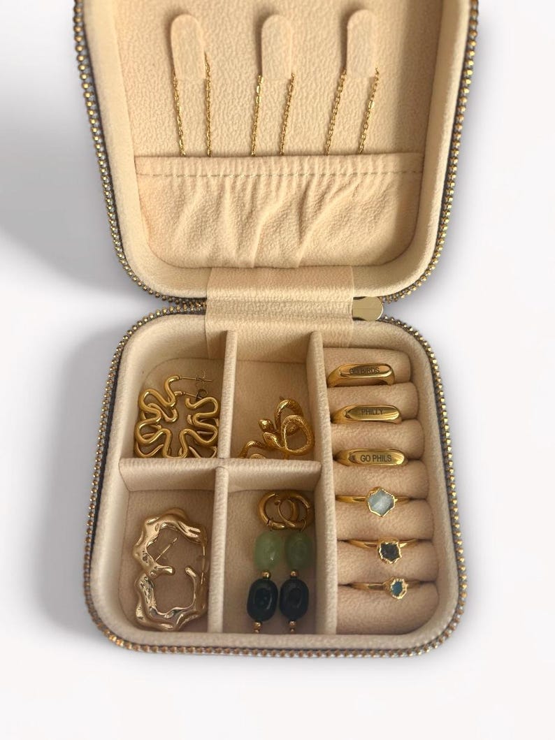 Pu&ograve; includere: Astuccio da viaggio per gioielli beige aperto, con vari orecchini, anelli e collane dorate. L'astuccio ha una chiusura a zip. Gli anelli includono il testo "GO PHILS". Orecchini con forme astratte e perline verdi e nere.