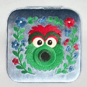 Pu&ograve; includere: Un specchietto compatto quadrato, blu chiaro, simile al velluto, con un ricamo di un volto di mostro verde e un bordo floreale. Il mostro ha grandi occhi bianchi, una cresta rossa e verde e una bocca nera. Il bordo floreale comprende fiori blu e rossi.