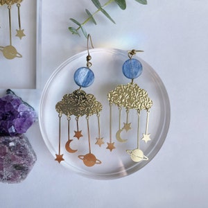 May include: A pair of dangle earrings featuring a blue circular bead attached to a textured gold-tone cloud. Hanging from each cloud are thin gold-tone metal chains with small star, crescent moon, and Saturn-shaped charms at the ends.
