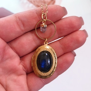 May include: Gold locket necklace with a blue stone and a small crystal charm. The locket is oval shaped and has a detailed design.