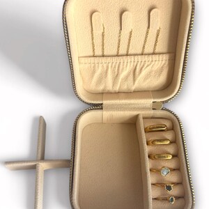 Pu&ograve; includere: Custodia da viaggio per gioielli beige aperta con cerniera e interno dorati. La custodia contiene orecchini a catena dorati e diversi anelli d'oro con pietre preziose. Gli anelli sono incisi con testo.