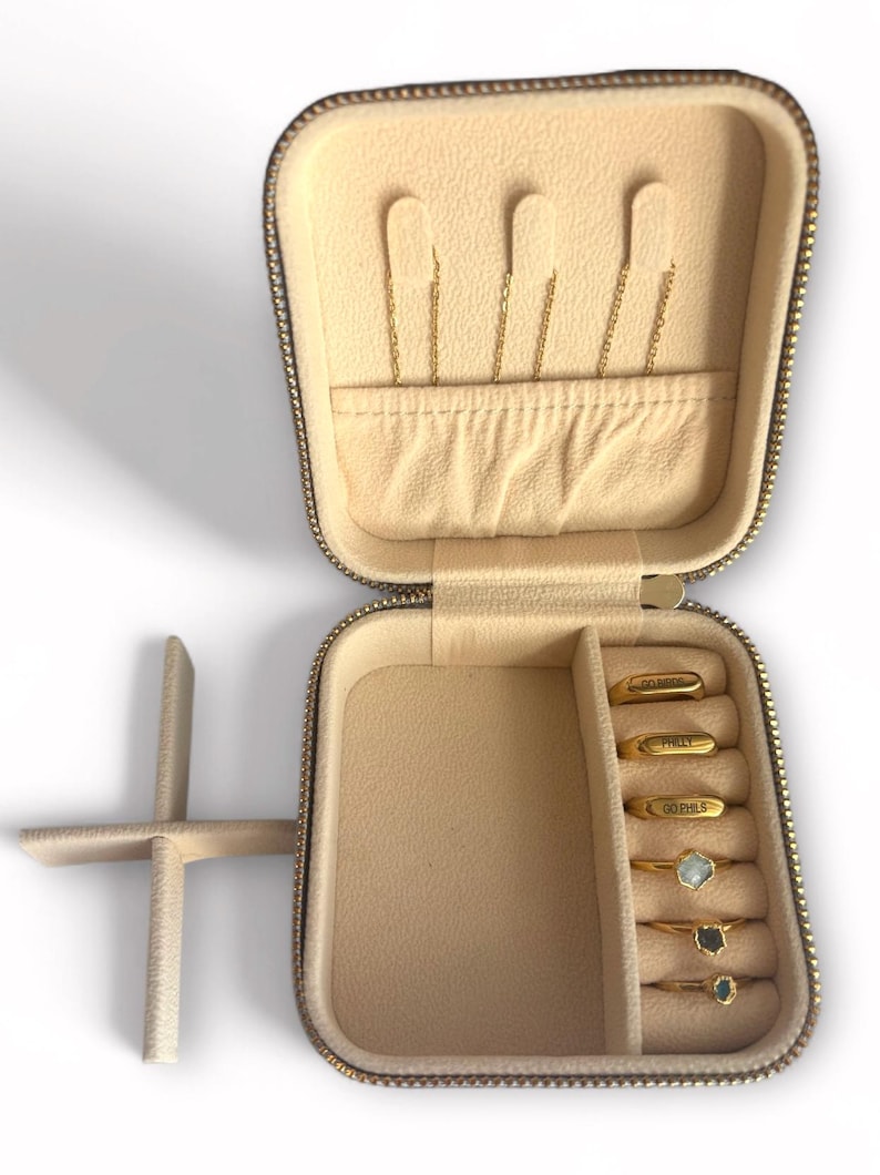 Pu&ograve; includere: Custodia da viaggio per gioielli beige aperta con cerniera e interno dorati. La custodia contiene orecchini a catena dorati e diversi anelli d'oro con pietre preziose. Gli anelli sono incisi con testo.