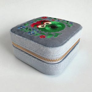 Pu&ograve; includere: Un portagioie quadrato grigio con cerniera dorata. La parte superiore &egrave; ricamata con un motivo floreale verde, rosso e blu. La scatola &egrave; realizzata in un materiale morbido e vellutato.