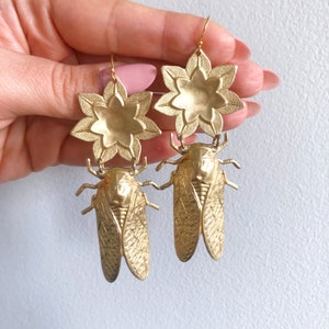 May include: Gold tone earrings featuring a cicada insect design with a flower charm above. The earrings are made of metal and have a textured finish.