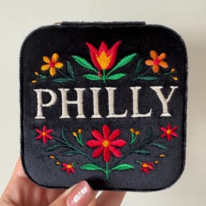 May include: Black, square jewellery case with embroidered floral design and the word "PHILLY" in white. The flowers are red, orange, and yellow with green leaves. The case is held by a hand.