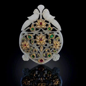 May include: A white carved stone pendant featuring two birds at the top and an intricate openwork floral design. The piece is inlaid with gold, small red rubies, and green emeralds, creating a detailed botanical pattern across the surface.
