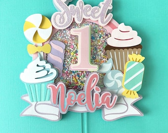 May include: A pink and white cake topper with the word "Sweet" and the number "1" in a pink circle with sprinkles. The name "Noelia" is written in pink below the number. The topper also features a cupcake, a lollipop, and candy.