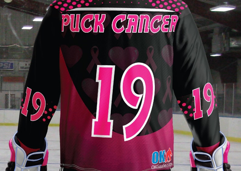 Puede incluir: Camiseta de hockey negra con detalles en rosa y las palabras "Puck Cancer" en la espalda. La camiseta tiene un n&uacute;mero rosa "19" en la espalda y corazones y cintas rosas en todo el dise&ntilde;o.