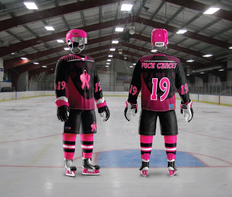 Puede incluir: Dos jugadores de hockey vistiendo jerseys rosas y negros con el texto "Puck Cancer" y el n&uacute;mero "19". Los jerseys tienen cintas rosas y corazones. Los jugadores est&aacute;n en una pista de hielo.