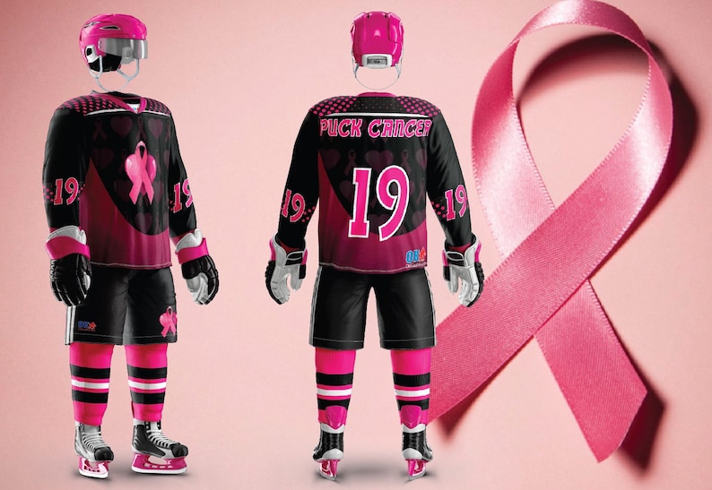 Puede incluir: Una camiseta de hockey negra con detalles rosas y un dise&ntilde;o de cinta rosa. La camiseta tiene el texto "Puck Cancer" y el n&uacute;mero "19" en la parte delantera. La camiseta la lleva un jugador de hockey con un casco rosa y guantes rosas.