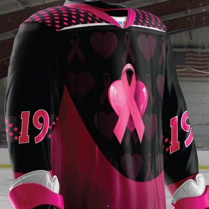 Puede incluir: Camiseta de hockey negra con corazones y cintas rosas. La camiseta tiene el n&uacute;mero 19 en ambas mangas.