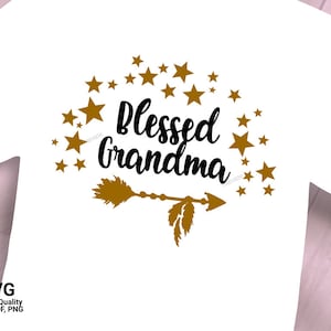 Puede incluir: Camiseta blanca con un diseño de flecha dorada y estrellas. El texto "Blessed Grandma" está escrito en negro.