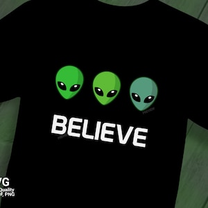 Op de afbeelding: Zwart T-shirt met drie groene alienhoofden en het woord "BELIEVE" in witte tekst.