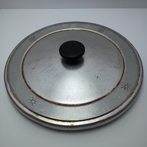 May include: A round, silver-colored metal pot lid featuring a central black knob. The surface shows signs of wear with visible scratches and light rust along the inner rim. Four small, engraved starburst patterns are spaced evenly around the lid.